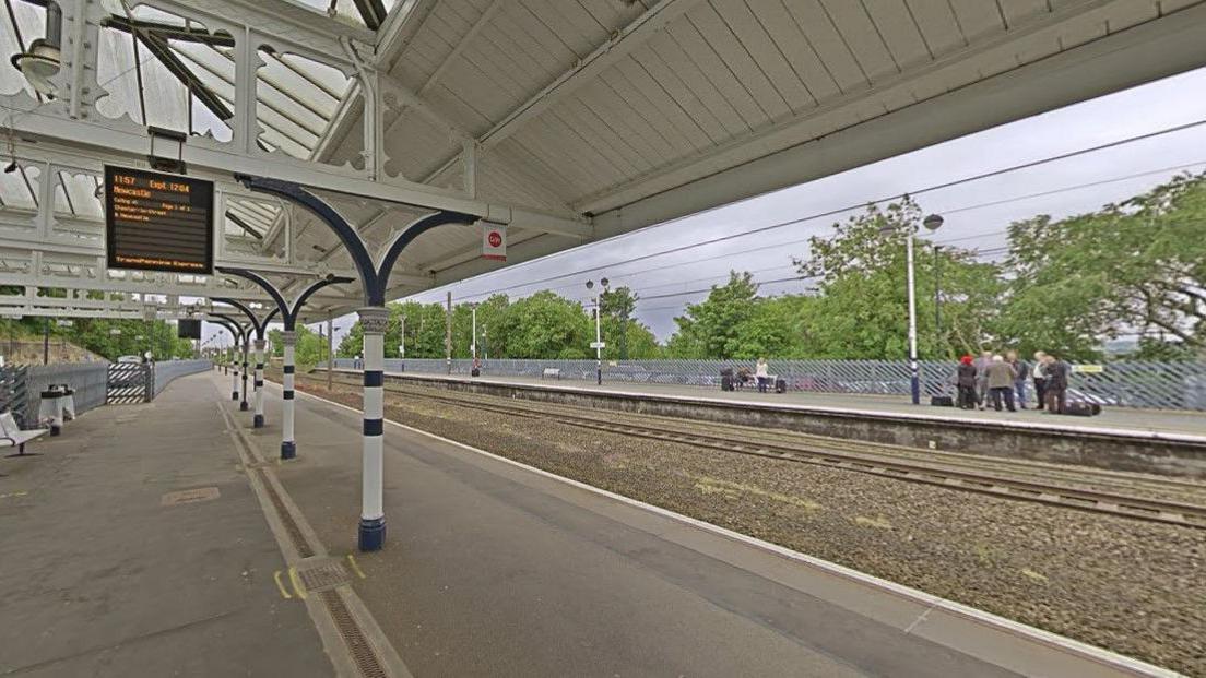 Northbound platform 2 at Durham station, overlooking the empty tracks. A screen is showing the next service to Newcastle. There are benches and bins. The roof of the platform is held by white and blue pillars.
A group of passengers is waiting on southbound platform 1 across the tracks. 