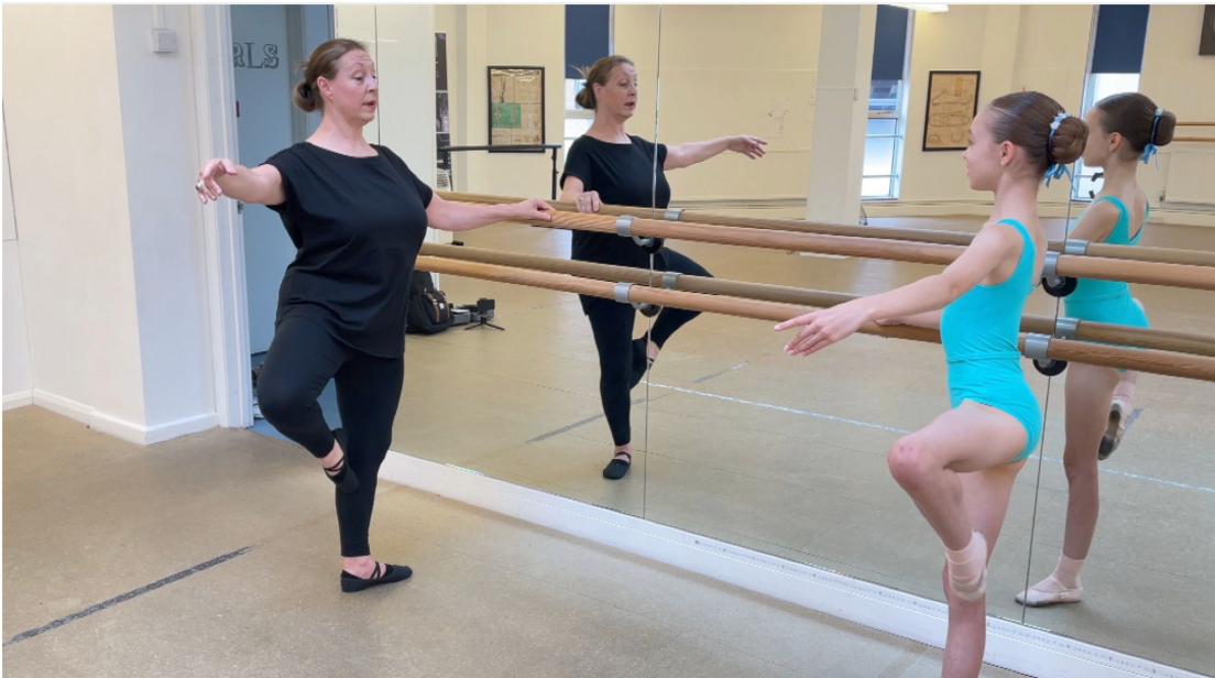 Trina Lee in black clothing doing a passé ballet, while holding on to the wooden bar attached to the mirror. Opposite is Jessica, in her blue leotard, mimicking the move.