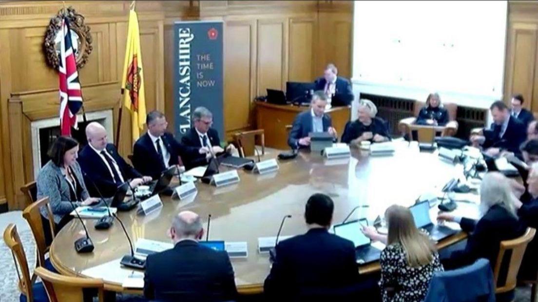 Councillors sit round an oval table - each has a laptop and stick mic in front of them.