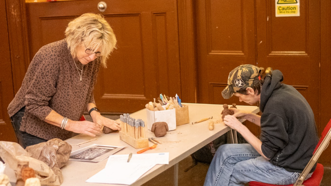 Bridget Hemmings working at a desk with a workshop participant sculpting his clay figure