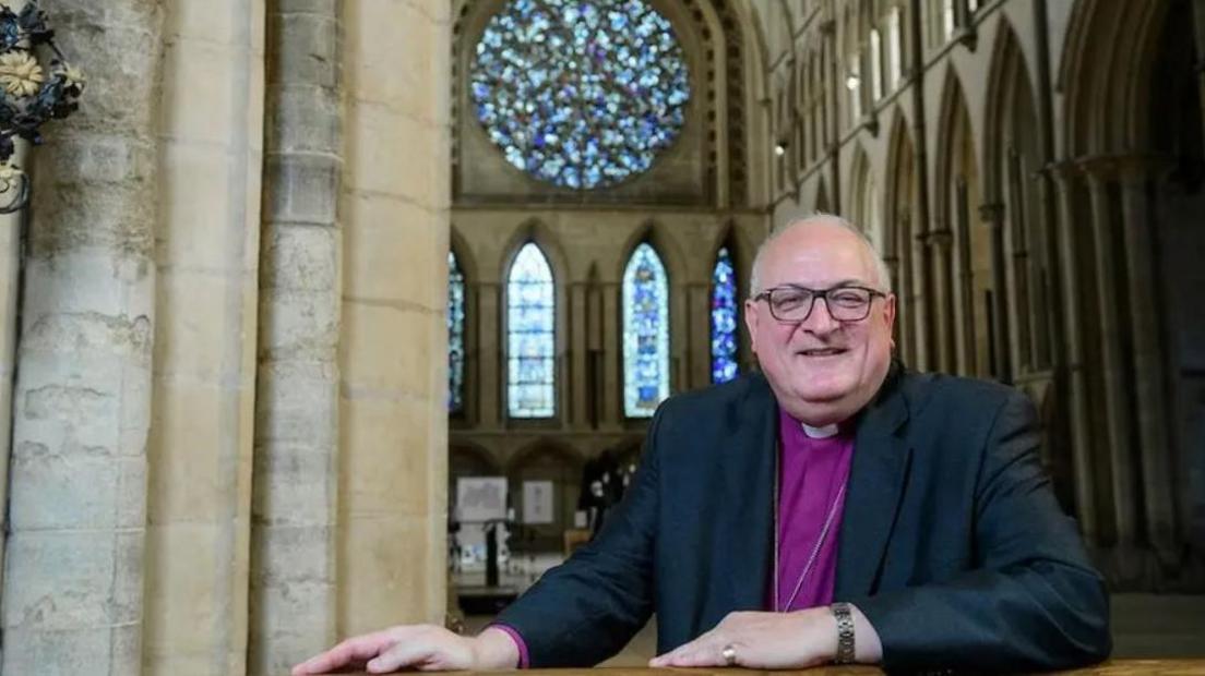 Bishop Stephen Conway, who is wearing glasses and is dressed in religious attire, sitting at a pew inside Lincolnshire Cathedral. The nave and a stained glass window can be seen behind him.