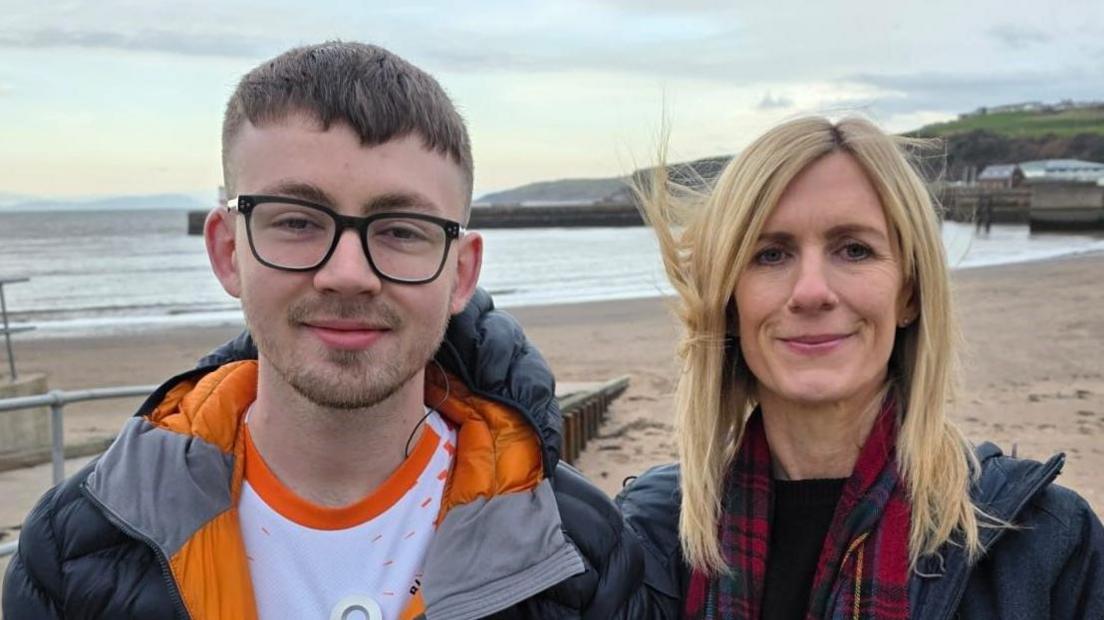 Adam Atkinson and Vicki Brown standing on a beach and smiling with the ocean in the background. Mr Atkinson has black-framed glasses, short cropped brown hair and stubble. He is wearing a black and orange jacket and a white and orange top. Ms Brown has blonde hair and is wearing a red scarf and a navy jacket.