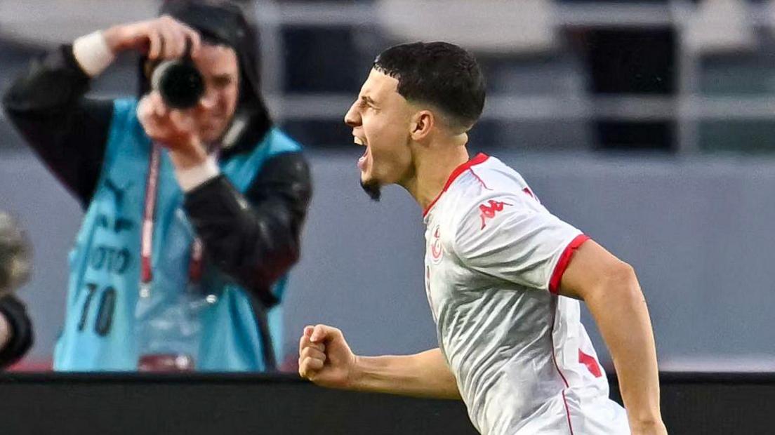 Ismael Garbi, wearing a white Tunisia shirt with red trim, screams in celebration and clenches his right fist after scoring for his country. A photographer in a light blue big takes his picture