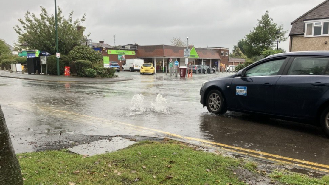 Water spurts up from a flooded road, as a taxi drives towards it. There is a Co-op supermarket in the distance, and the sky is grey