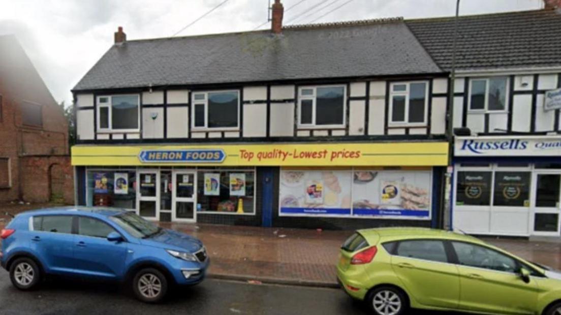 Heron Foods is in a mock Tudor row of terraced houses and has a yellow shop front with a blue sign. It has been raining and two cars are parked in front of the shop.