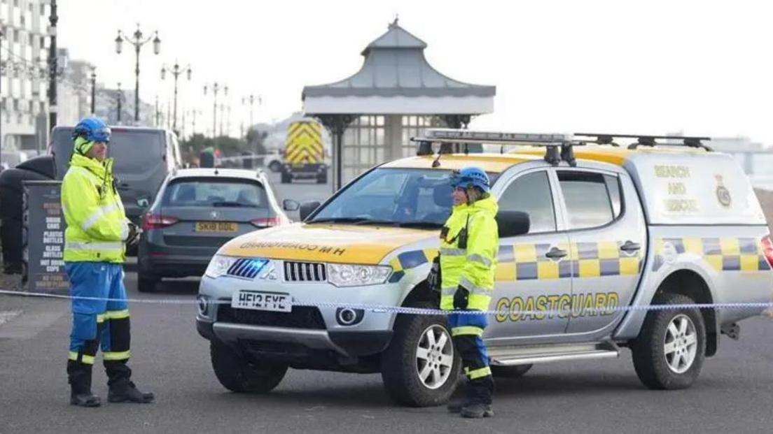 Two coastguard officers in hi vis outfits in front of a taped off area and their vehicle on Worthing seafront