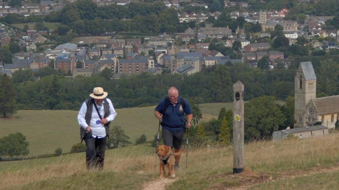 Robin Shuckburgh and former RAF Warrant Officer Al Sylvester MBE during the walk. The village of Ryeford, Gloucestershire, is behind them. They are smiling as they walk. Mr Shuckburgh is holding his dog on a leash. It's a sunny day.