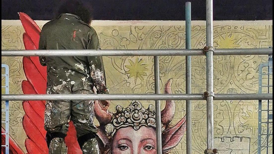 A street artist in khaki overalls spattered with paint standing on scaffolding and painting the side of a building with a large mural of a dragon with a woman's head