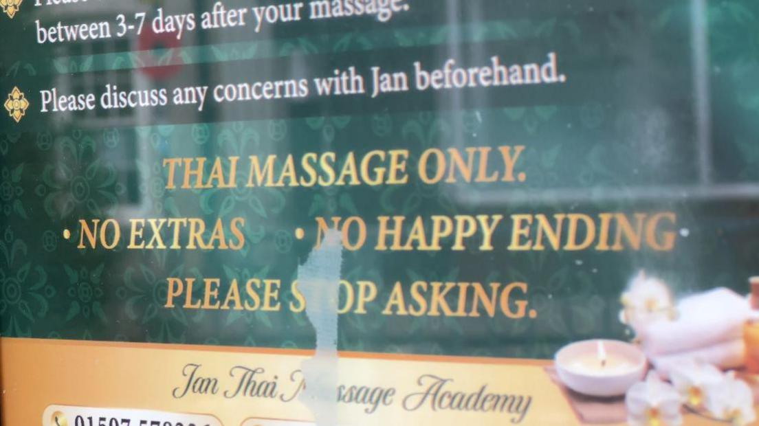 A zoomed-in view of a sign on Ms Raithby's shop. The text is yellow on a green background and reads: "Thai massage only, no extras, no happy ending, please stop asking." The sign also has an image of candles and flowers.
