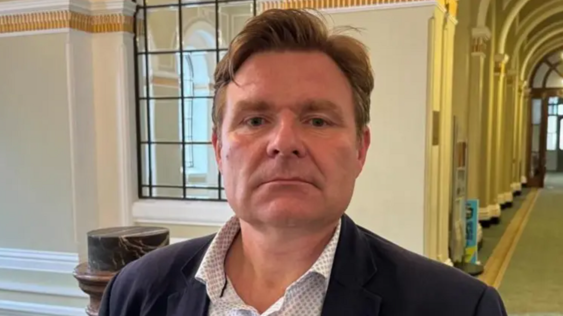 Council leader John Cotton pictured with a blank expression on his face. He has light brown hair which is styled in a quiff. He is wearing a dotted white shirt underneath a blue navy blazer