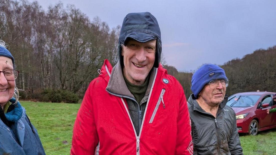 A man in a red coat and a black hat smiling at the camera with a field and a row of trees behind him.