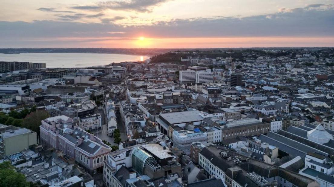 St Helier's skyline with thousands of buildings dotted around as the sun sets in the horizon over the sea.