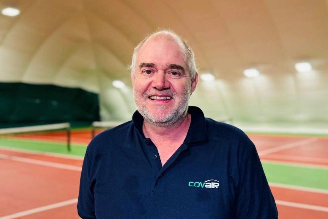 Peter Brown on a tennis court inside a dome, smiling at camera.