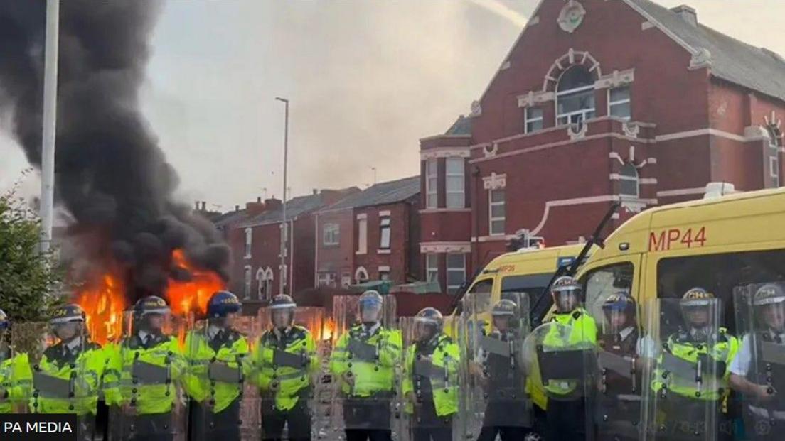 Line of riot police in high vis clothing holding riot shields outside mosque where one police van is on fire 