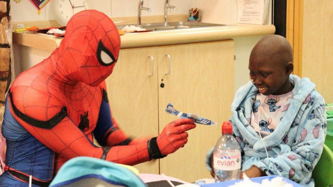 Spiderman with 10-year-old Jasmine from Peterborough. Spiderman is playing with a toy plane as Jasmine sits next to him and smiles. Jasmine wears a 'Lilo and Stitch' blue dressing gown with matching pyjamas. She has no hair and a thin, soft tube is attached to her nose.
