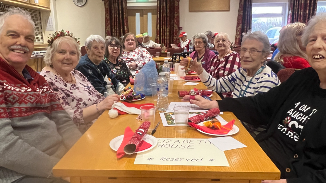 A group of pensioners round a festive table in a care home, all smiling with crackers on plates in front of them.