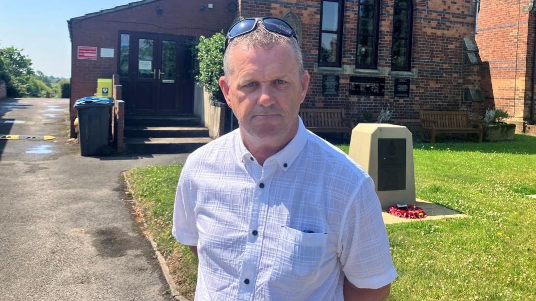 The image shows a man standing in front of a red-brick church. He is wearing a short-sleeved white button-up shirt. Sunglasses are resting on top his head.