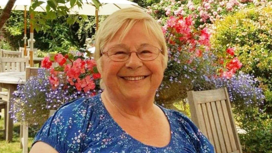 Gwynneth Exall smiles at the camera. She is wearing a blue t-shirt with small white and pink flowers and has glasses. She is sitting in a garden on wooden garden furniture with flowers in the background.