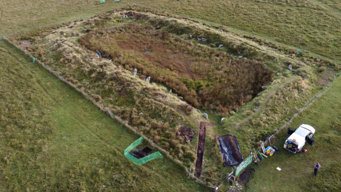 An aerial shot of a rectangular mound on moorland. The mound has an inner rectangle of granite standing stones, and an archeological trench in one corner.