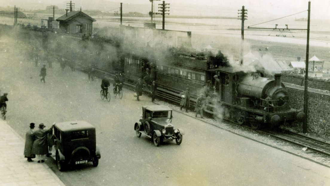 A black and white photo of a steam locomotive steaming along a seaside line with old cars on the road in the foreground and a beach and sea in the background