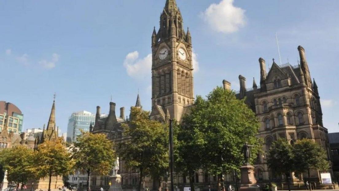 Manchester Town Hall seen behind a row of trees, with the clock tower visible, along with modern and historic buildings nearby.