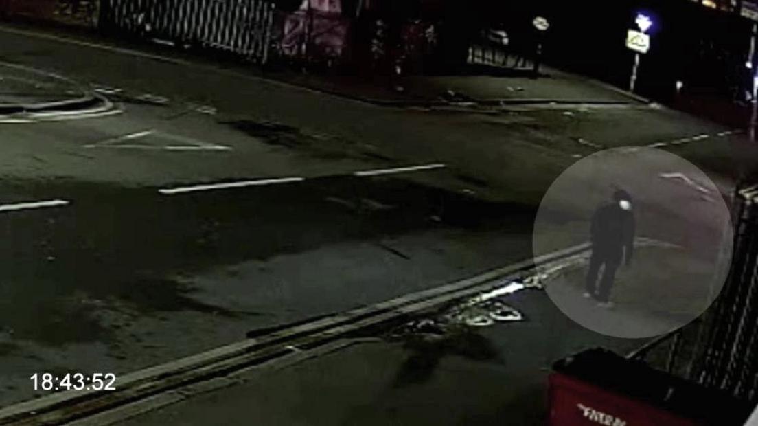 An image captured on CCTV of the killer. He is walking on the pavement and is wearing dark clothing. You cannot make out his face. It is evening time and a road can be seen behind him but there is no sign of any cars. Only a street sign can be seen lit up in the background.