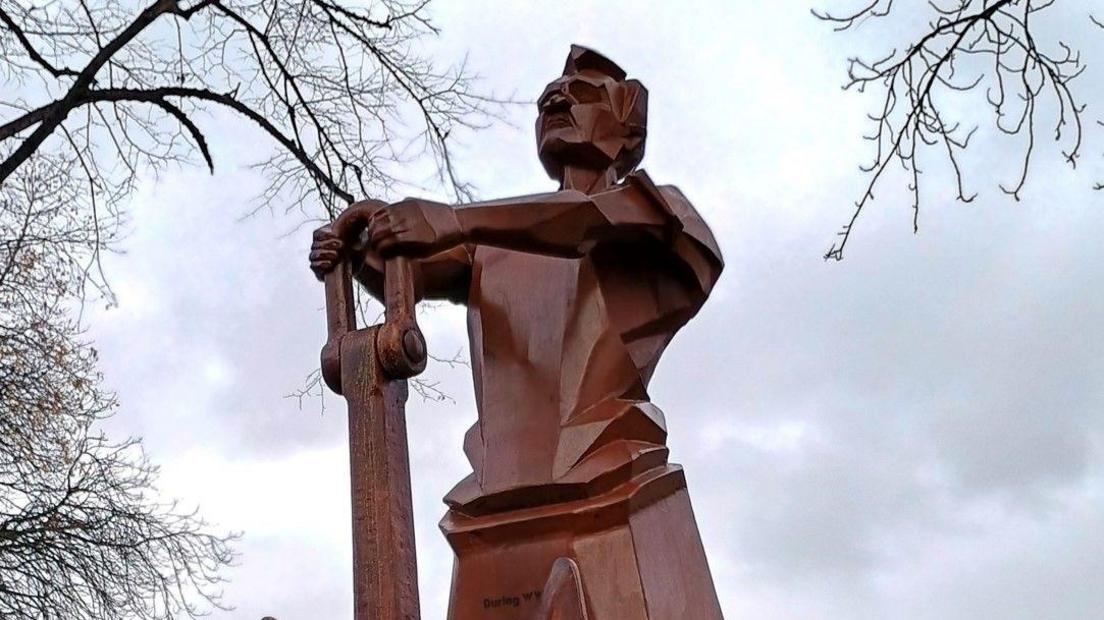 A brown-coloured monument depicting a man, who was in a reserved occupation during World War Two. Trees and sky are in the background.