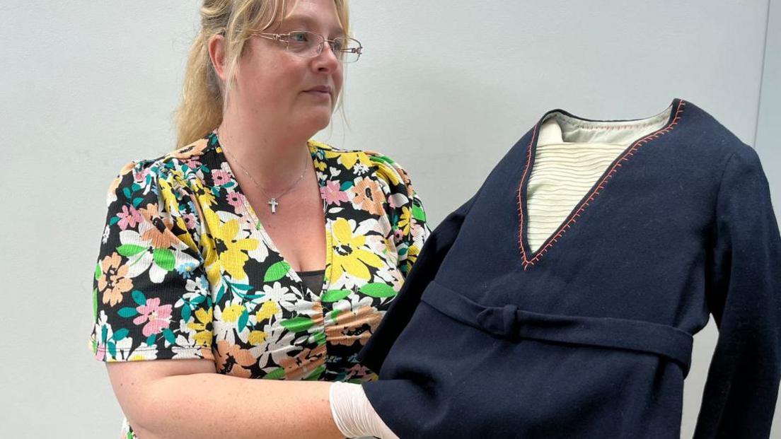 A woman wearing glasses and a floral dress holds up a children's navy blue sailor suit top.