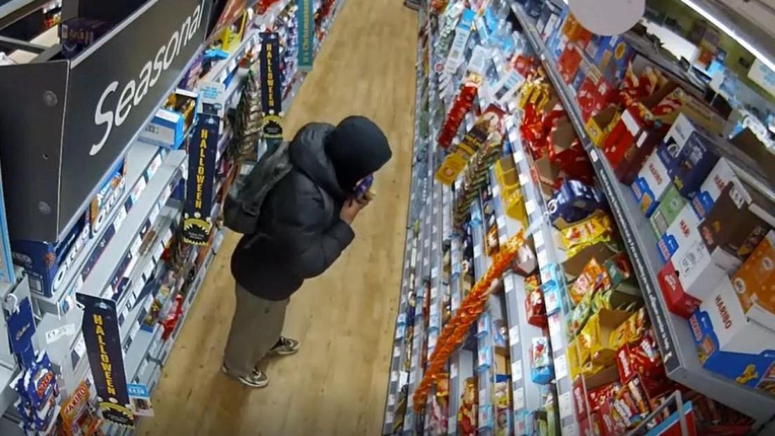 Man with a dark puffer jacket and hoodie puts chocolate into his jacket in a convenience store aisle.