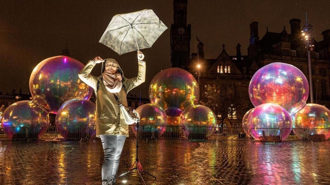 Several large inflatable bubbles featuring as part of an art installation in City Park - a public square in Bradford. A woman is stood in front of the installation wearing a rain jacket with her hood up, jeans and is holding an umbrella in the air. 