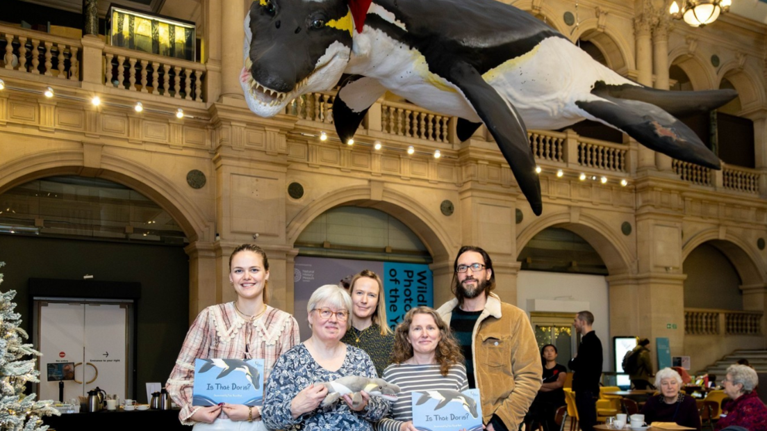 A group of people hold the book 'Is That Doris?' while stood underneath a model of Doris the pliosaur.