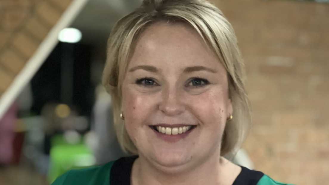 A woman smiling at the camera, she has short blonde hair and is wearing a green and black dress. 