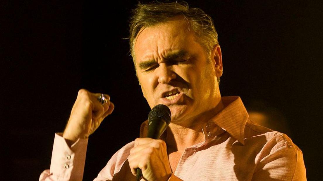 Morrissey, wearing a pink open-necked shirt with short side-parted hair and his eyes closed, holds one hand in a fist while singing into a microphone held in the other