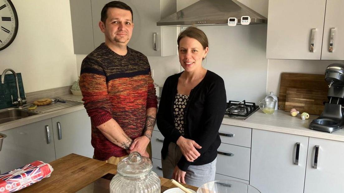 A man wearing a jumper is on the left next to a woman with dark hair wearing a dark top. They are in the kitchen looking at the camera. Cupboards are behind them and a table, with items on, is in front of them.