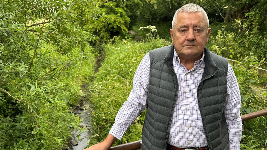Gavin Heskett, wearing a chequered white and blue shirt and a grey gilet, stands in front of the dene which he believes caused his pet Jasper to become unwell and die.
