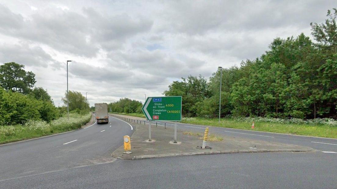 A road junction with a green sign in the middle pointing to Stoke-on-Trent, Congleton and Crewe. There are trees on embankments either side of the road