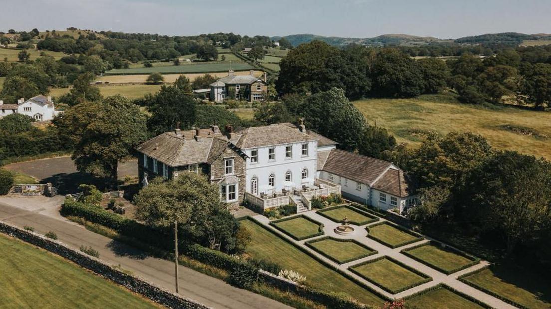 An aerial shot of the old grammar school building in Cartmel. The large white building is L-shaped and sits next to a stone-clad one. There is a large manicured garden at the front.