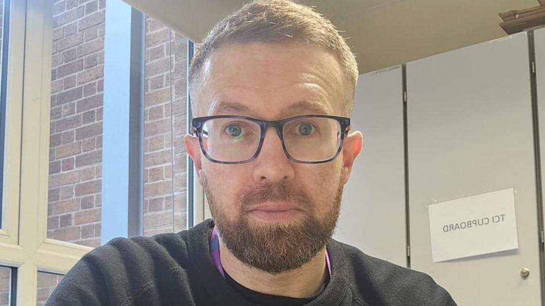 Darragh Statham sits in front of a window and grey cupboard. He is wearing a black jumper and glasses. He has brown hair and a beard.