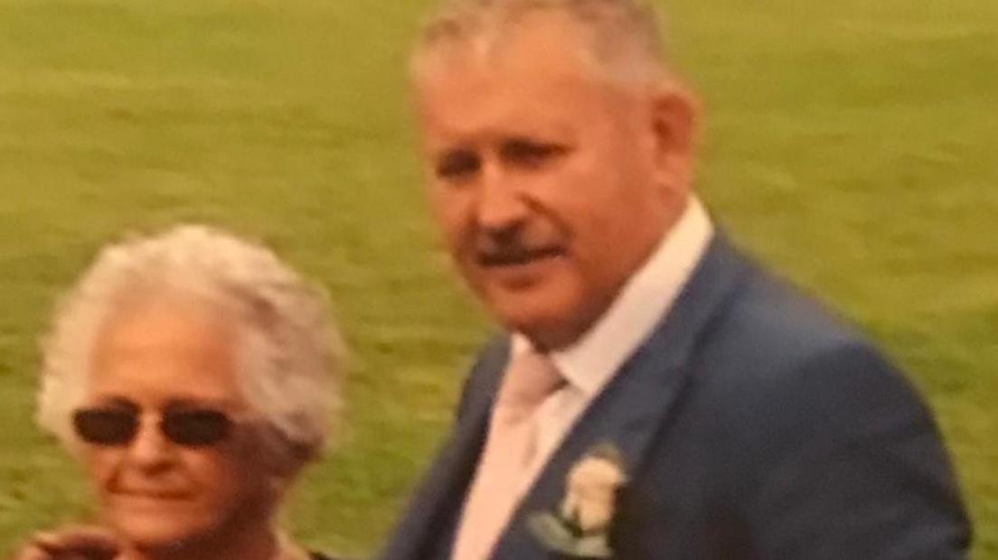 A man with grey hair and a grey moustache wearing a navy blue wedding suit, stood next to a woman with short grey hair and oval shaped sunglasses