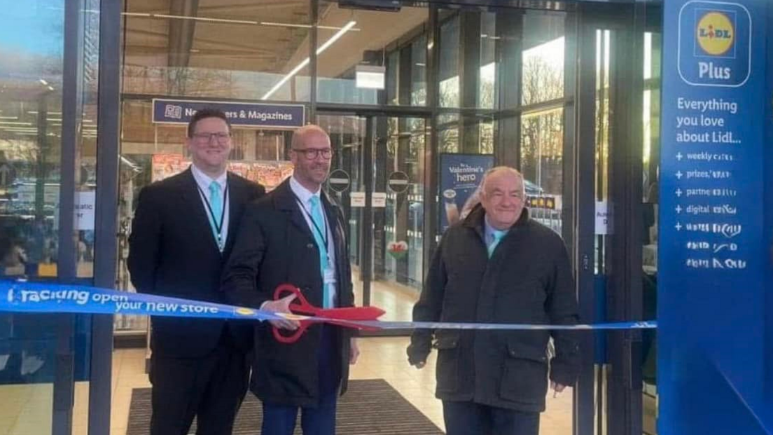 The three Reform councillors all wearing black jackets, white shirts and pale blue ties. Two are bald, while the third has short dark hair. The one in the middle is holding a pair of giant red scissors as they stand behind a Lidl branded ribbon which has the words 'new store' on it, with the glass doors of Lidl in Cwmbran behind them.