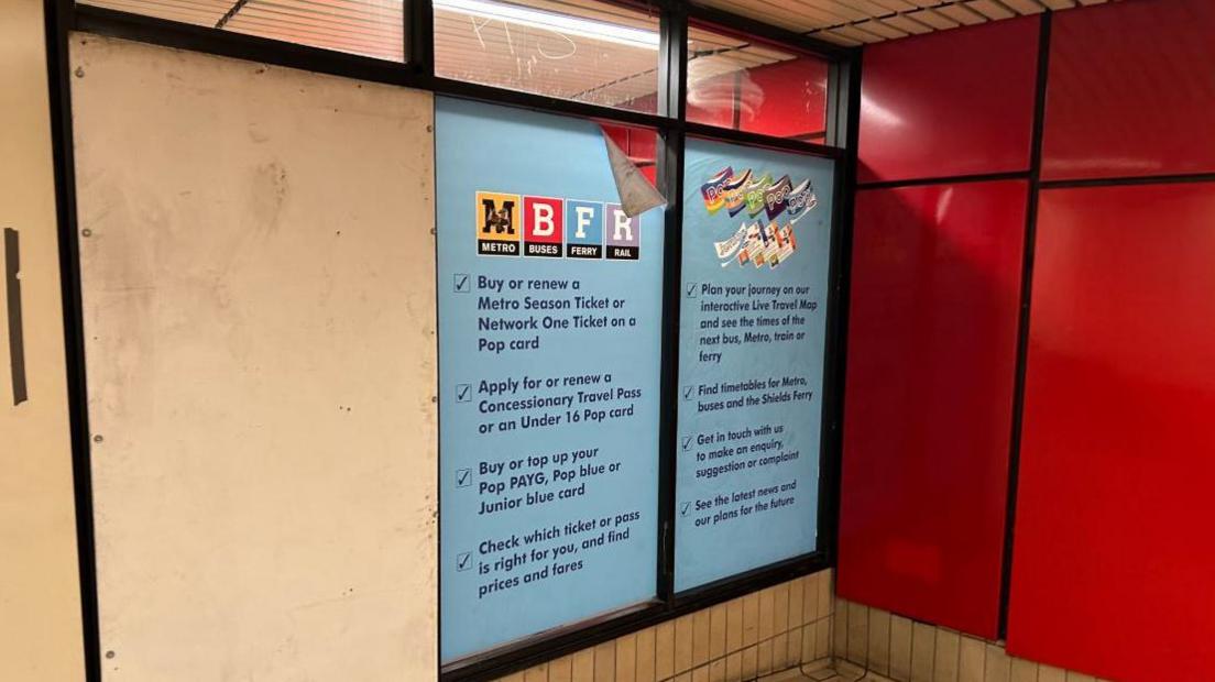 View of the entrance to a former Metro travel office. A boarded up door is to the left, next to two blue panels with information about tickets and passes, and narrow windows above. To the left, at right angles, is a wall with red panels.