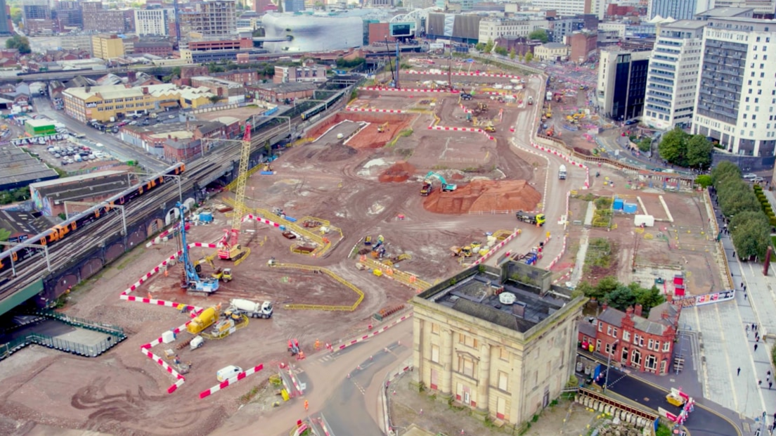 An aerial shot of the construction site. Flat land can be seen with various coned off, or barricaded areas, with some cranes.