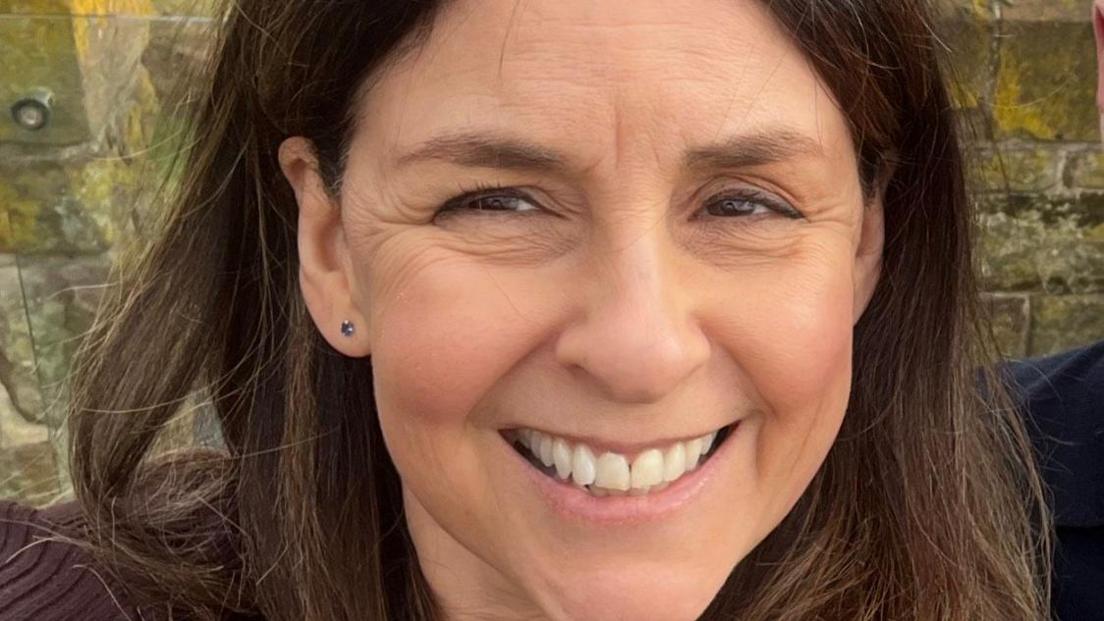 Hilary Mitchell is smiling at the camera. She has long brown hair and is wearing studded earrings.