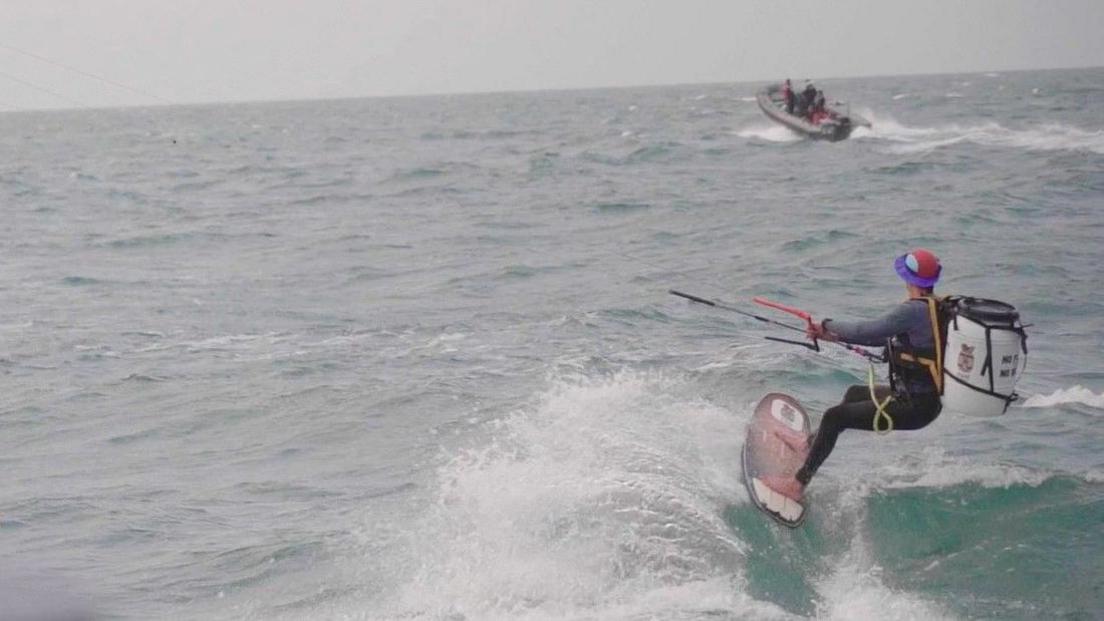 Martin Marescaux kitesurfing on the sea with a large white barrel on his back. There is a small RIB boat in the background.