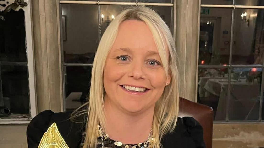 April Jeffers smiles facing the camera as she sits in a room with stone walls and large windows. She has straight, blonde hair below her shoulders, wears a black dress or top and holds a golden trophy from an awards ceremony.