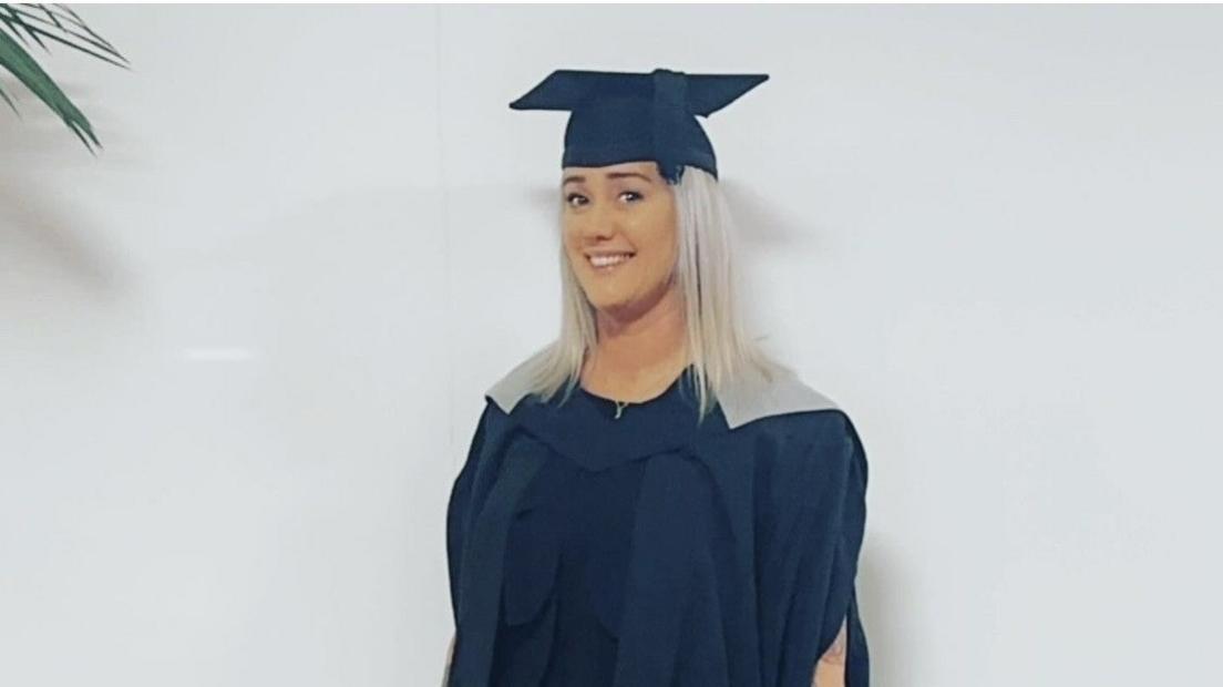 A woman with blonde hair poses in a university graduation hat and cape.