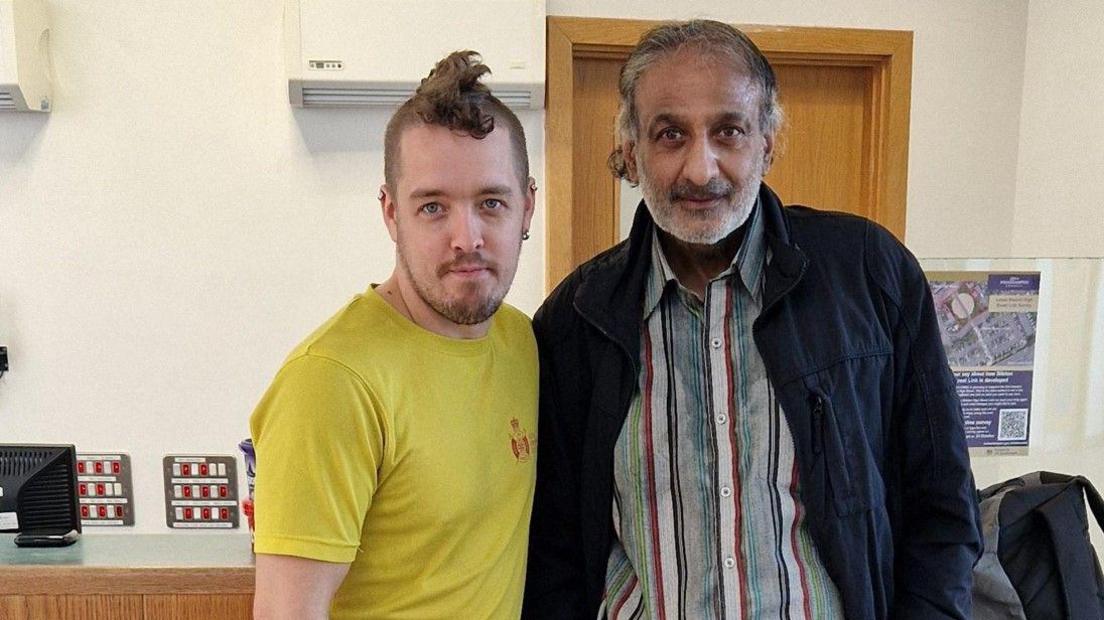 A man with brown hair in a yellow T-shirt on the left is next to an older man in a dark jacket and striped shirt. A door is behind him.