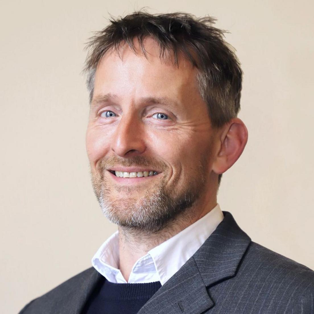 A man with grey-ish brown hair and a beard in a jacket with a white shirt and blue jumper underneath.