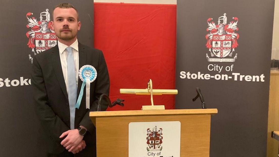 A man stands next to a podium at a by-election count. He has his hands clasped in front of him and he's wearing a blue and white Reform rosette.
