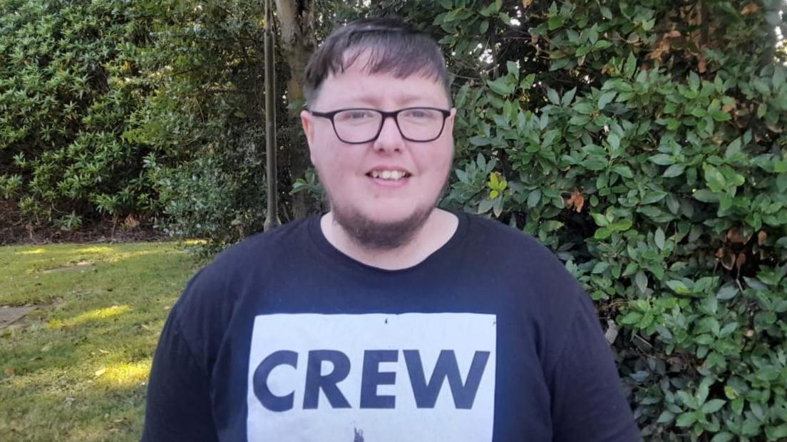 Jason Dunne is standing in a black T-shirt with the word 'Crew' visible in a white box. He is wearing glasses and has stubble. He is standing near to a grassy area with a number of trees visible.